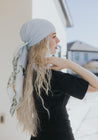 Woman with long wavy blonde hair wearing a light blue silk headscarf tied with a flowing white panel featuring painted bird and foliage designs, standing outdoors with soft natural lighting.