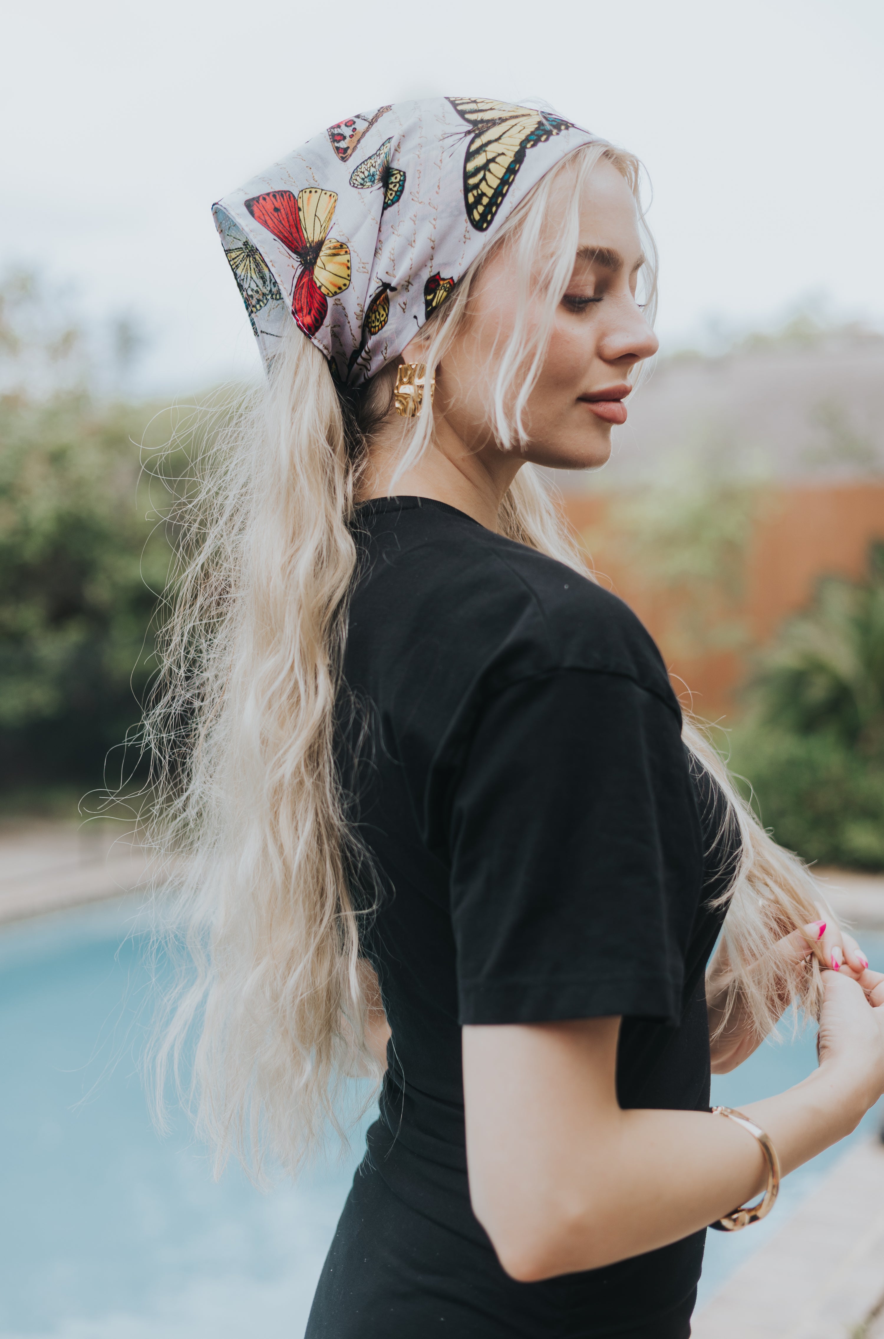 Woman wearing a white silk headscarf with a colorful butterfly print, styled in a retro wrap over long wavy blonde hair, standing poolside in an outdoor setting.